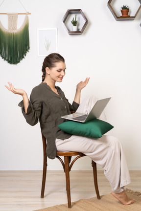 Vista di tre quarti di una giovane donna che indossa abiti da casa seduta su una sedia con un laptop e alzando le mani