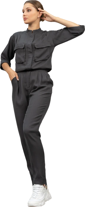 Front view of a walking young woman in a jumpsuit holding hand in pocket & touching head  download