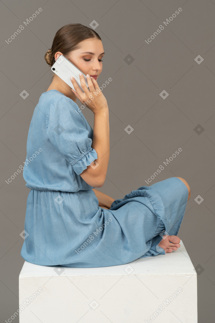 Seitenansicht einer jungen frau, die auf einem würfel sitzt und mit dem smartphone spricht
