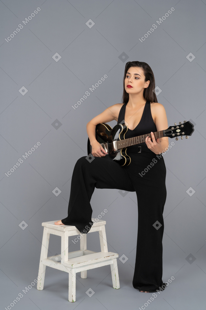 Beautiful young woman playing a black guitar while keeping her leg on a stool