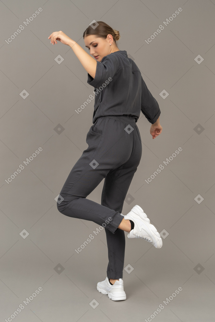 Three-quarter back view of a young woman in a jumpsuit raising hand and leg