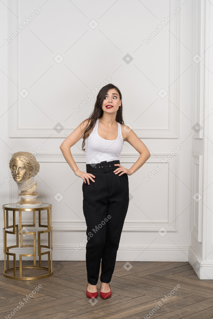 Front view of a perplexed young female putting hands on hips while standing near golden greek sculpture
