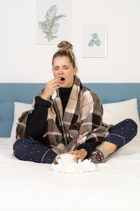 Vista frontal de una joven enferma bostezo en pijama envuelto en una manta marcada en la cama
