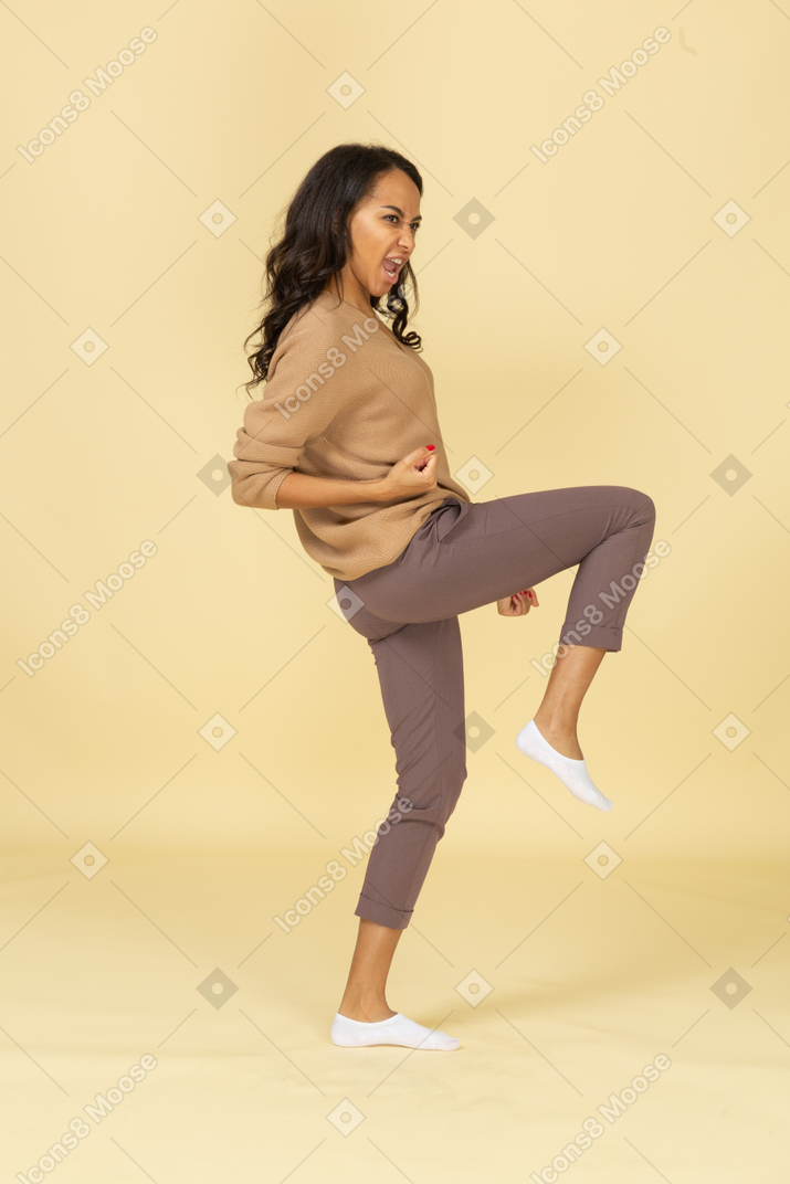 Vista lateral de una fresca mujer joven de piel oscura levantando la pierna y apretando el puño