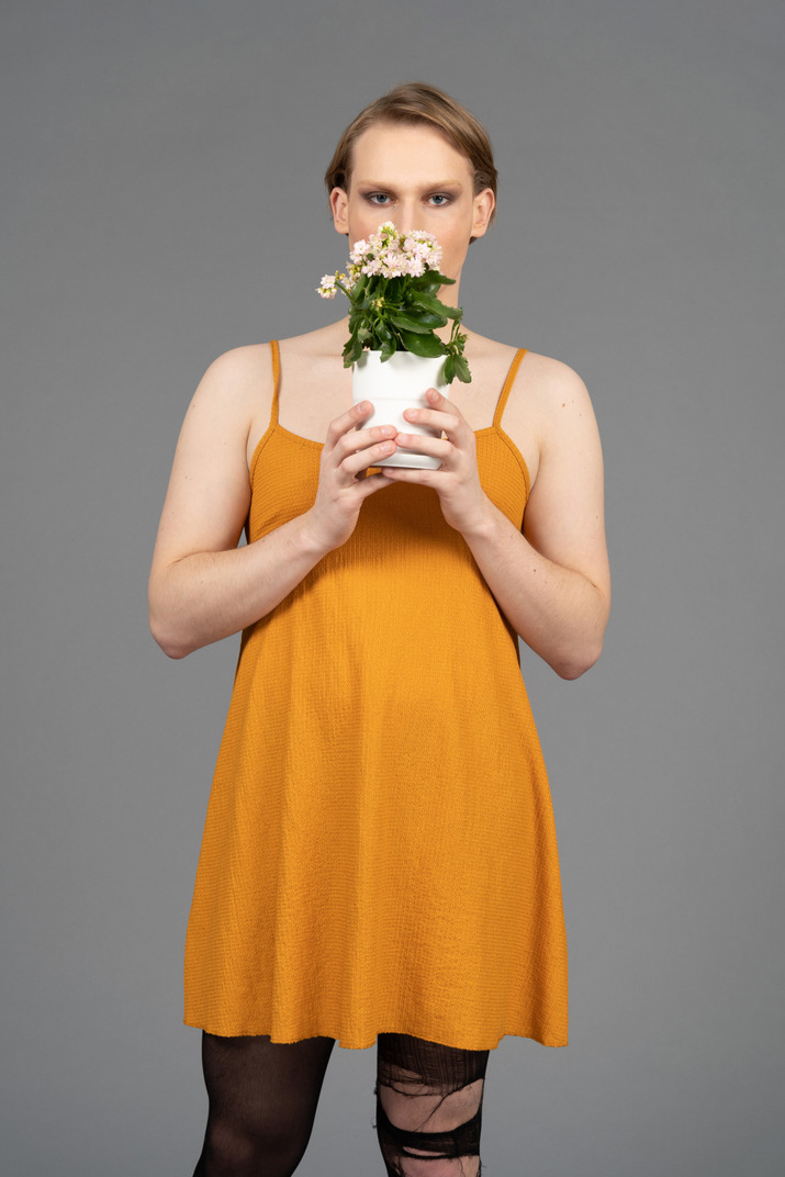 Front view of a young queer person in orange dress smelling flowers