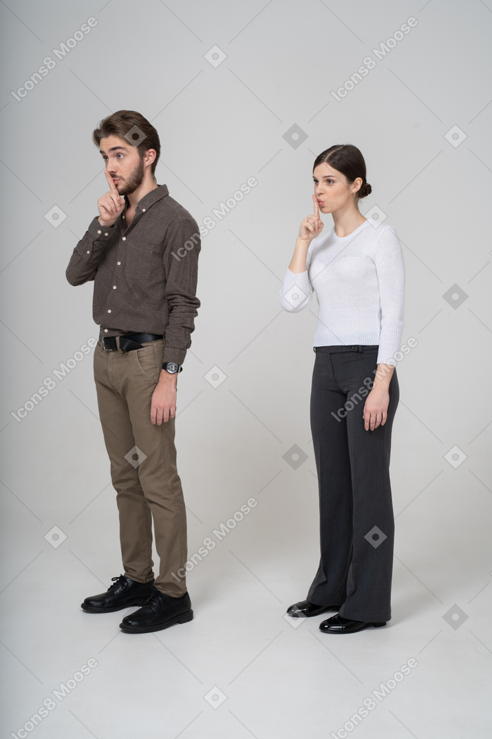 Vista de três quartos de um jovem casal com roupas de escritório mostrando gesto de silêncio