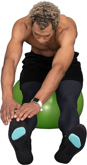 一个赤膊的非洲人坐在绿色健身球上伸展的前视图