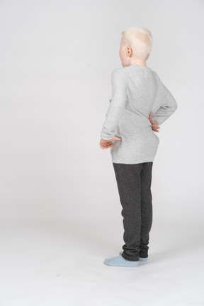 Vista posterior de tres cuartos de un niño chico solitario en ropa casual poniendo la mano en la cadera