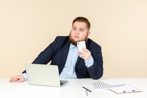 Pensive young overweight man sitting at the office desk and holding phone