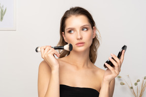 Front view of a young woman applying face powder while holding a mirror