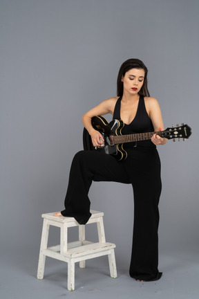 Vista frontal de una joven en mono negro sosteniendo la guitarra y poniendo la pierna en el taburete