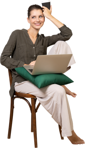 Front view of a young woman wearing home clothes sitting on a chair with a laptop and coffee