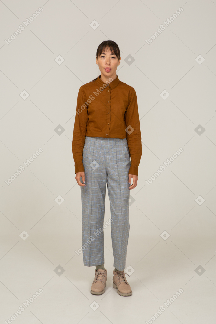 Vista frontale di una giovane donna asiatica in calzoni e camicetta che incrocia le braccia e mostra la lingua