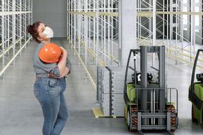 Plump masked woman standing in a warehouse and holding protective helmet
