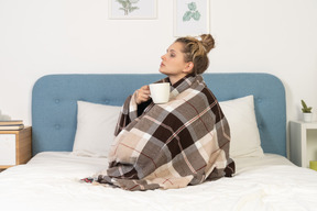 Vista lateral de una joven enferma envuelta en una manta marcada en la cama sosteniendo una taza de té