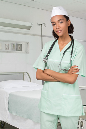 Young female doctor standing with her hands crossed near the hospital bed