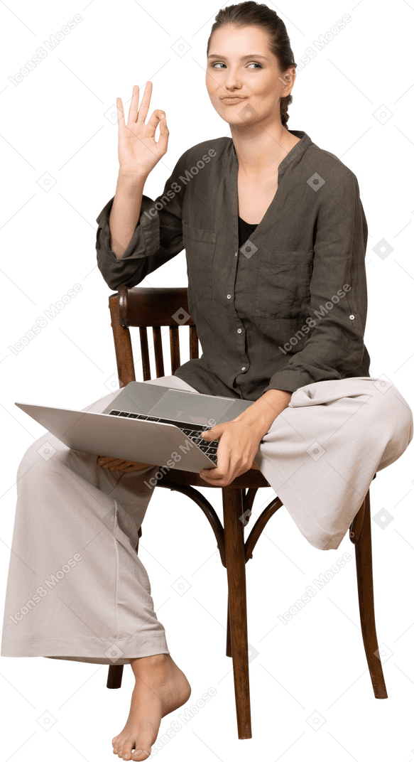 Front view of a funny young woman sitting on a chair with a laptop & showing ok gesture