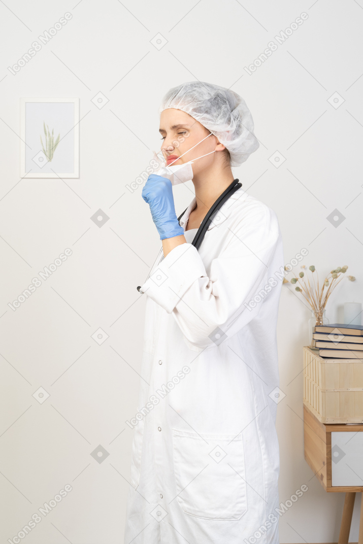 Vista de tres cuartos de una joven doctora disgustada tratando de quitarse la máscara