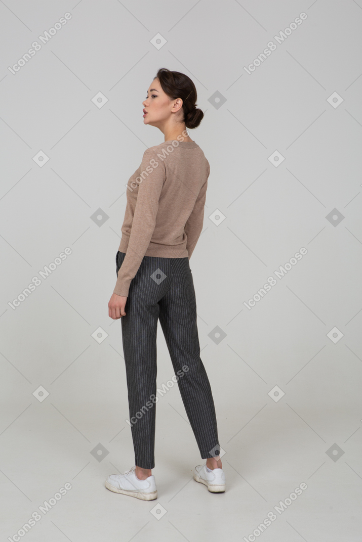 Vista posterior de tres cuartos de una señorita parada quieta en suéter y pantalones mirando a un lado