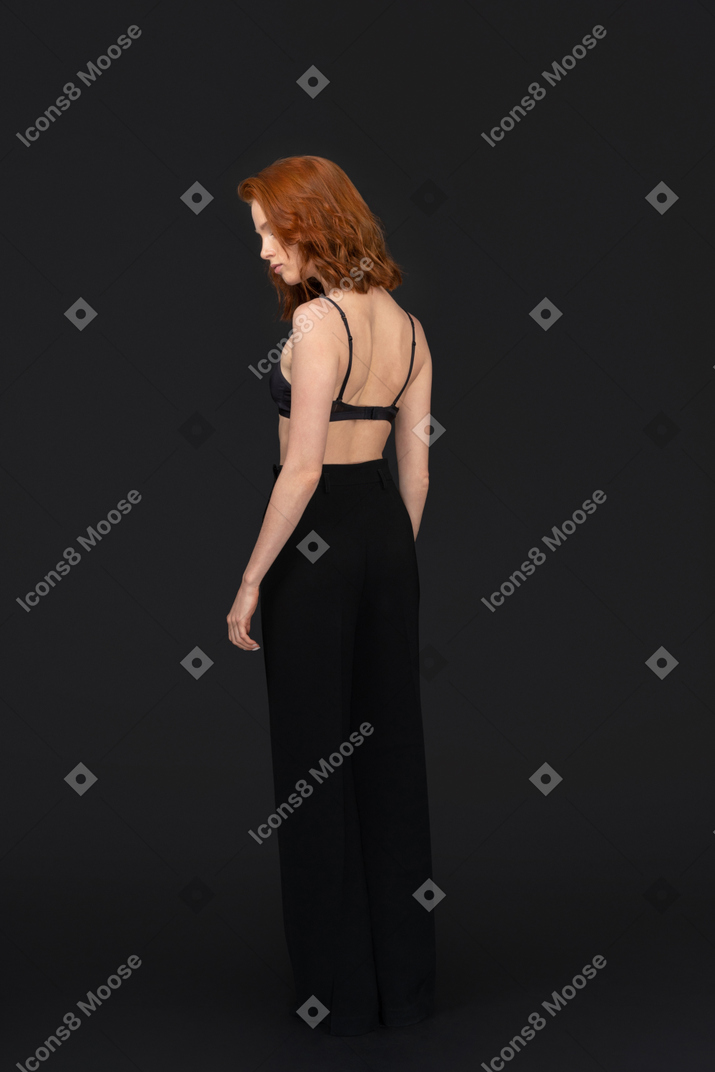 A three-quarter back side view of the sexy young woman standing on the black background