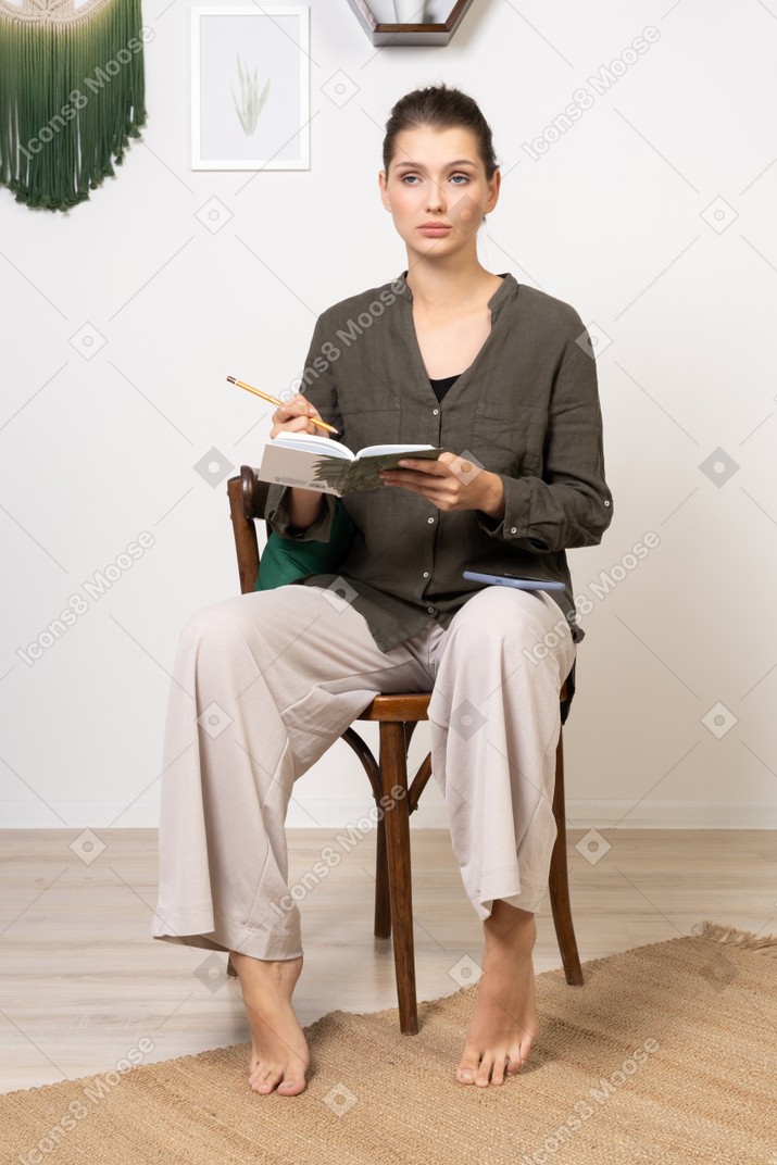 Vista frontale di una giovane donna premurosa che indossa abiti da casa seduta su una sedia con matita e taccuino