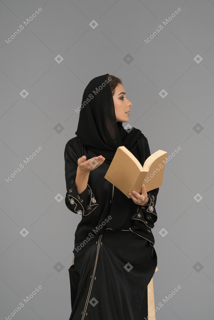 A gesturing woman with a book looking sideways