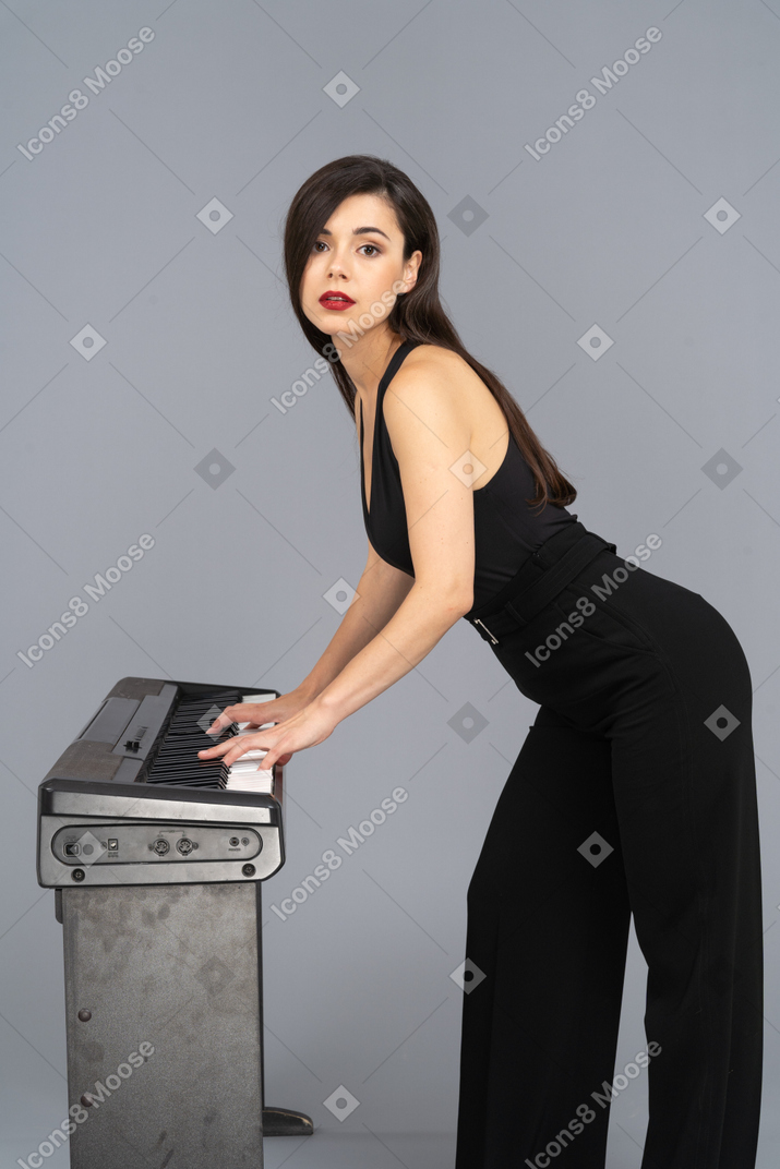 Side view of a young lady in black suit playing the piano while raising head while looking at camera