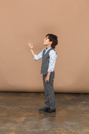 Vista lateral de um menino de terno cinza mostrando sinal de ok