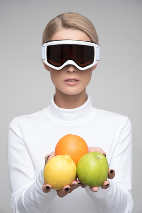 Jovem loira com óculos de esqui segurando frutas coloridas