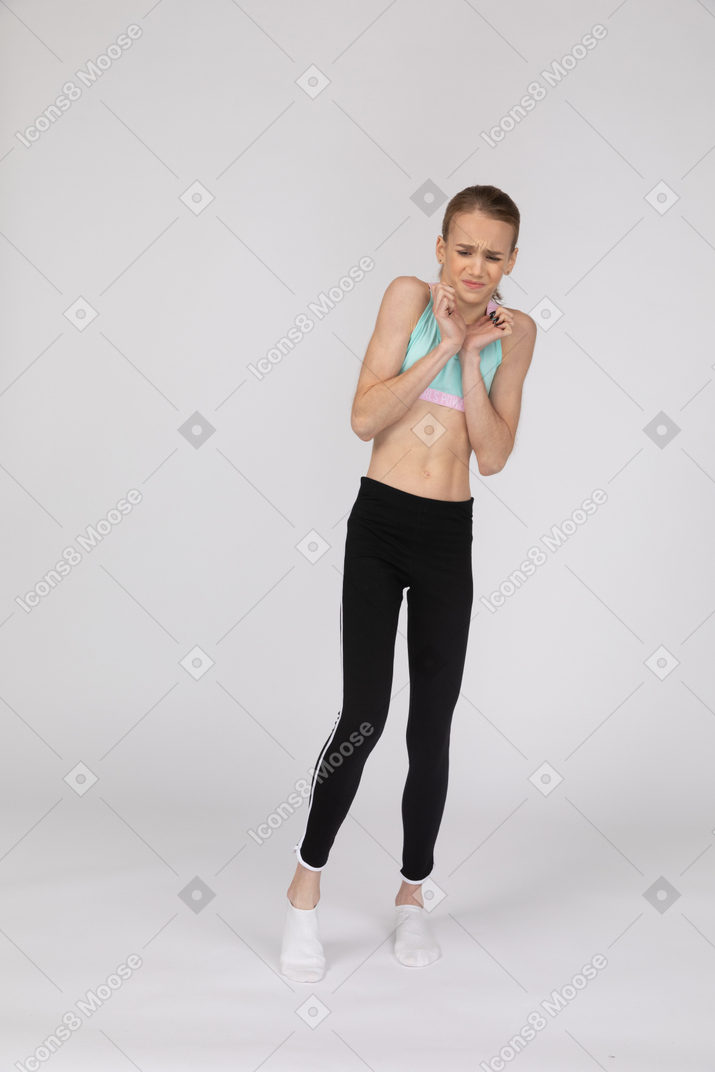 Front view of a displeased teen girl in sportswear narrowing eyes and holding hands together