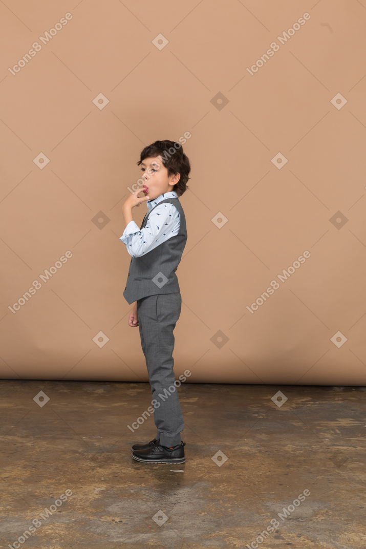 Vista lateral de um menino de terno cinza lambendo o dedo