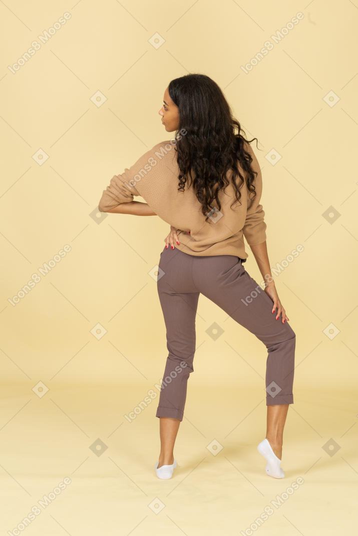 Back view of a dark-skinned young lady putting hand on hip while looking aside