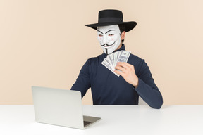 Hacker wearing vendetta mask sitting at the table and counting money