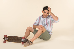 Joven caucásico ajustando el cabello y sentado cerca de skate