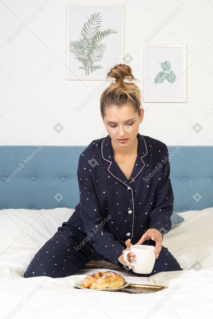 Vista frontal de una joven en pijama sosteniendo una taza de café y algunos pasteles en una bandeja