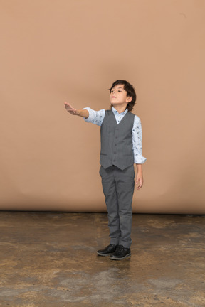 Vista frontal de un chico lindo con traje gris de pie con el brazo extendido