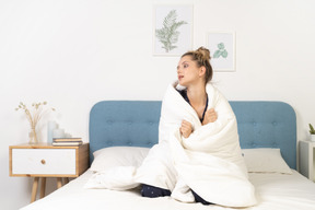 Front view of a tired young woman in pajamas wrapped in blanket staying in bed