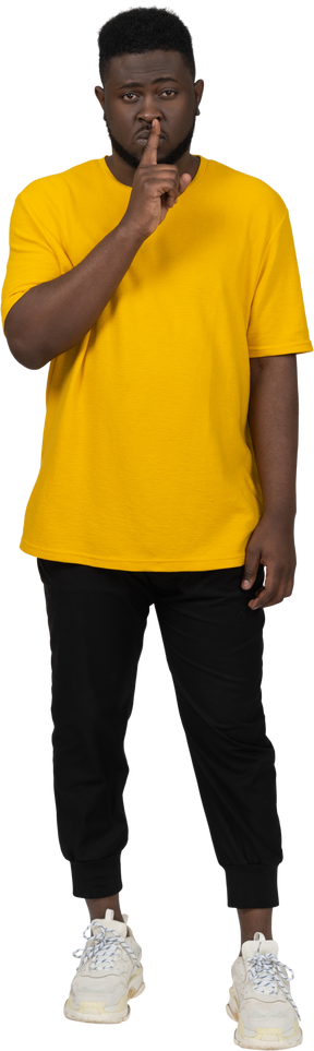 Vista frontale di un giovane uomo dalla pelle scura in maglietta gialla che mostra gesto di silenzio