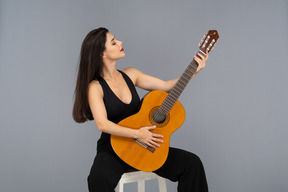 Front view of a sitting young lady in black suit looking proudly at her guitar