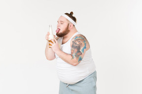 一个胖胖的运动员拿着一瓶啤酒，伸出舌头