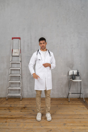 Front view of a young doctor standing in a room with ladder and chair showing a size of something