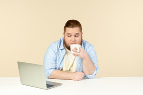 Молодой человек с избыточным весом пьет чай и смотрит что-то на ноутбуке