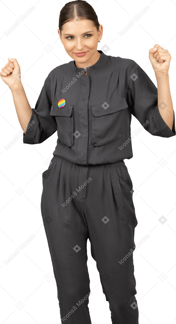 Front view of a young woman in a jumpsuit with lgbt pin raising hands & clenching fists