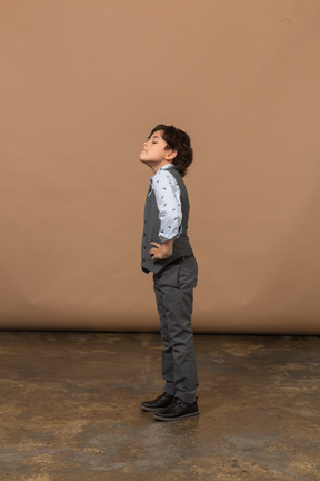 Vista laterale di un ragazzo carino in abito grigio in piedi con le mani sui fianchi e alzando lo sguardo