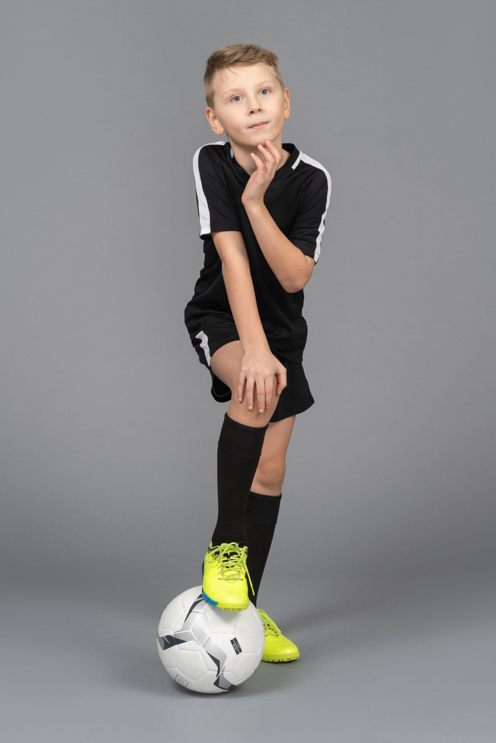 Front view of a child boy in football uniform putting his foot on ball and touching chin