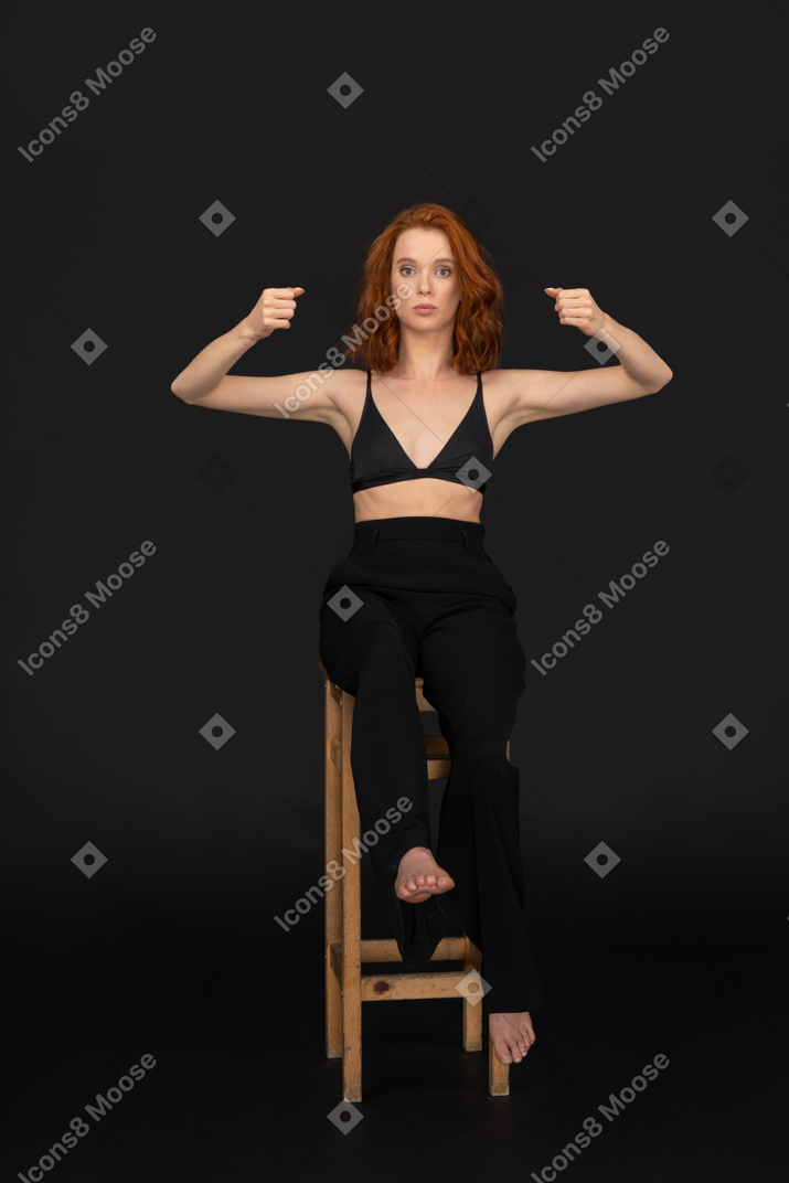 A frontal view of the beautiful woman dressed in black pants and bra, balancing on the chair