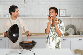 A man cooking meat on a grill and standing next to a woman eating avocado
