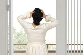 A woman in a white dress holding her head and looking out a window