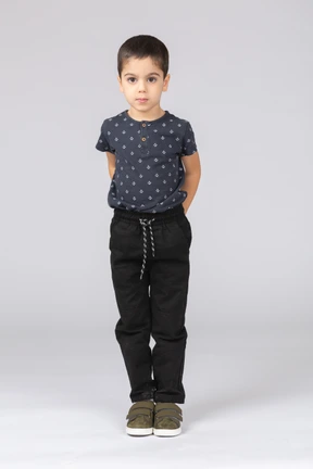 Front view of a cute boy standing with hands behind back and looking at camera