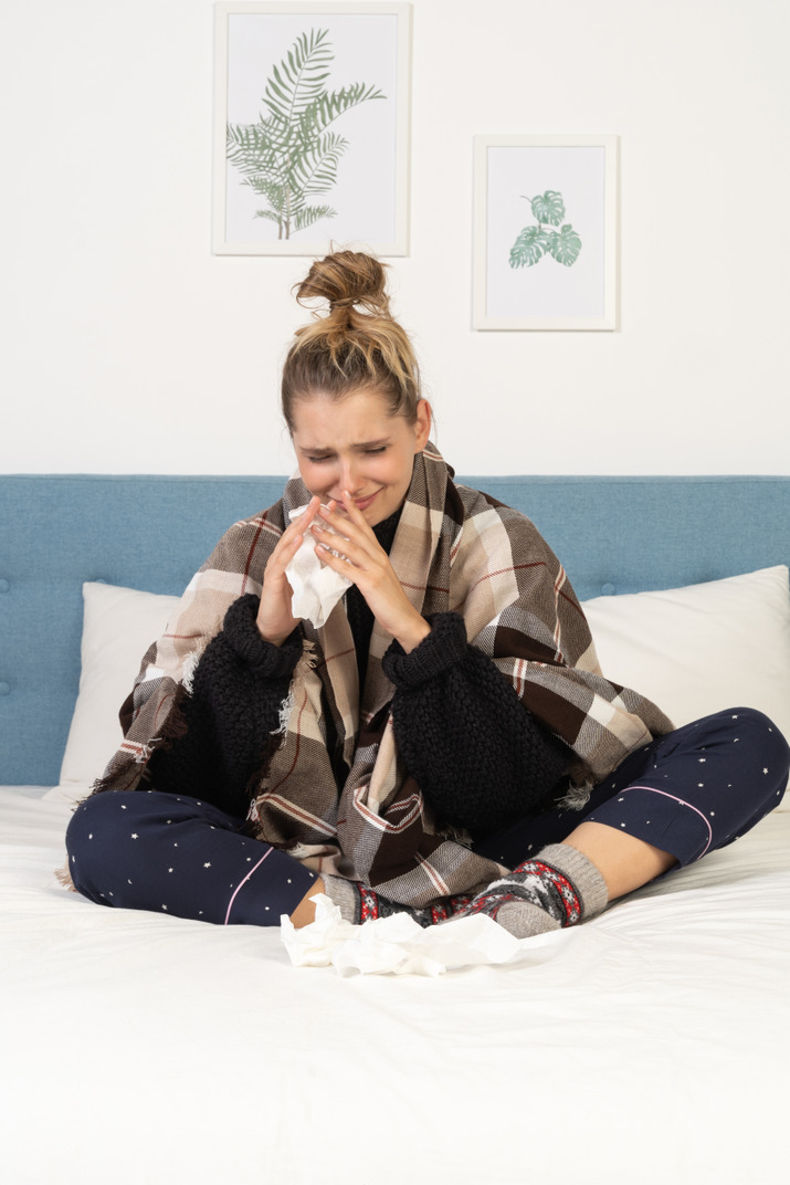 Front view of an ill young lady in pajamas wrapped in checked blanket in bed blowing her nose